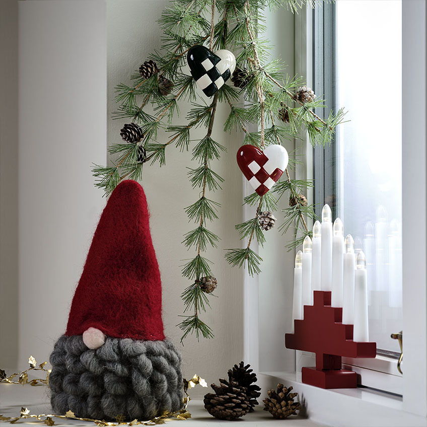 Lutin de Noël avec chapeau de lutin rouge et barbe grise, arc de bougie rouge et brindille de sapin artificiel.