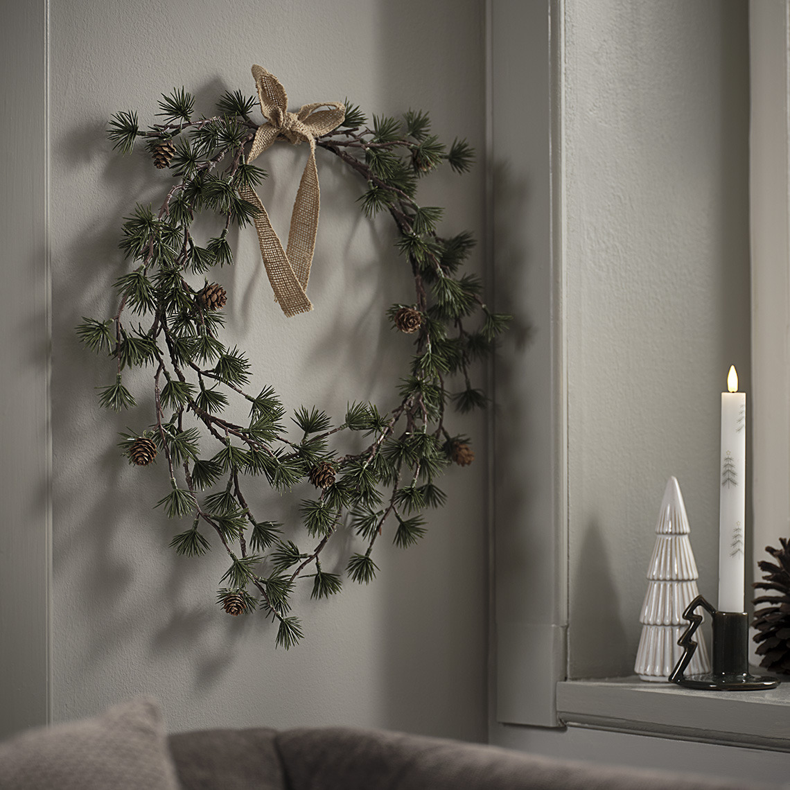 Couronne de Noël BELI, arbre de Noël décoratif MYSING et bougeoir STILBIT avec bougie de Noël STILBIT Couronne de Noël suspendue près d’un rebord de fenêtre avec un arbre décoratif de Nöel et une bougie de Noël