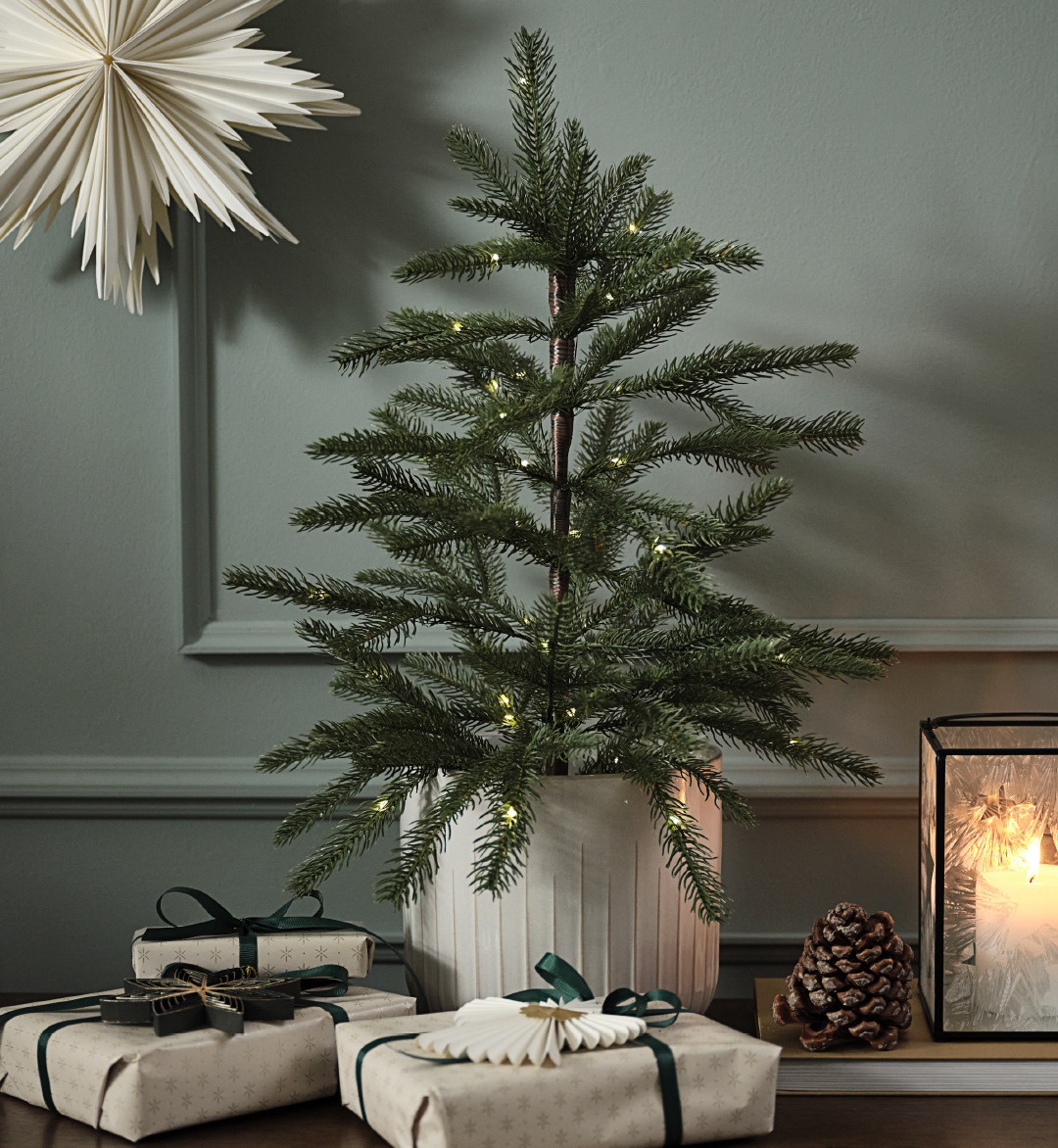Kersboom BERLINGR, lantaarn OLIVIN, inpakpapier MAGNESIT Kleine kerstboom tegen groene muur met wanddecoratie en ingepakte cadeautjes
