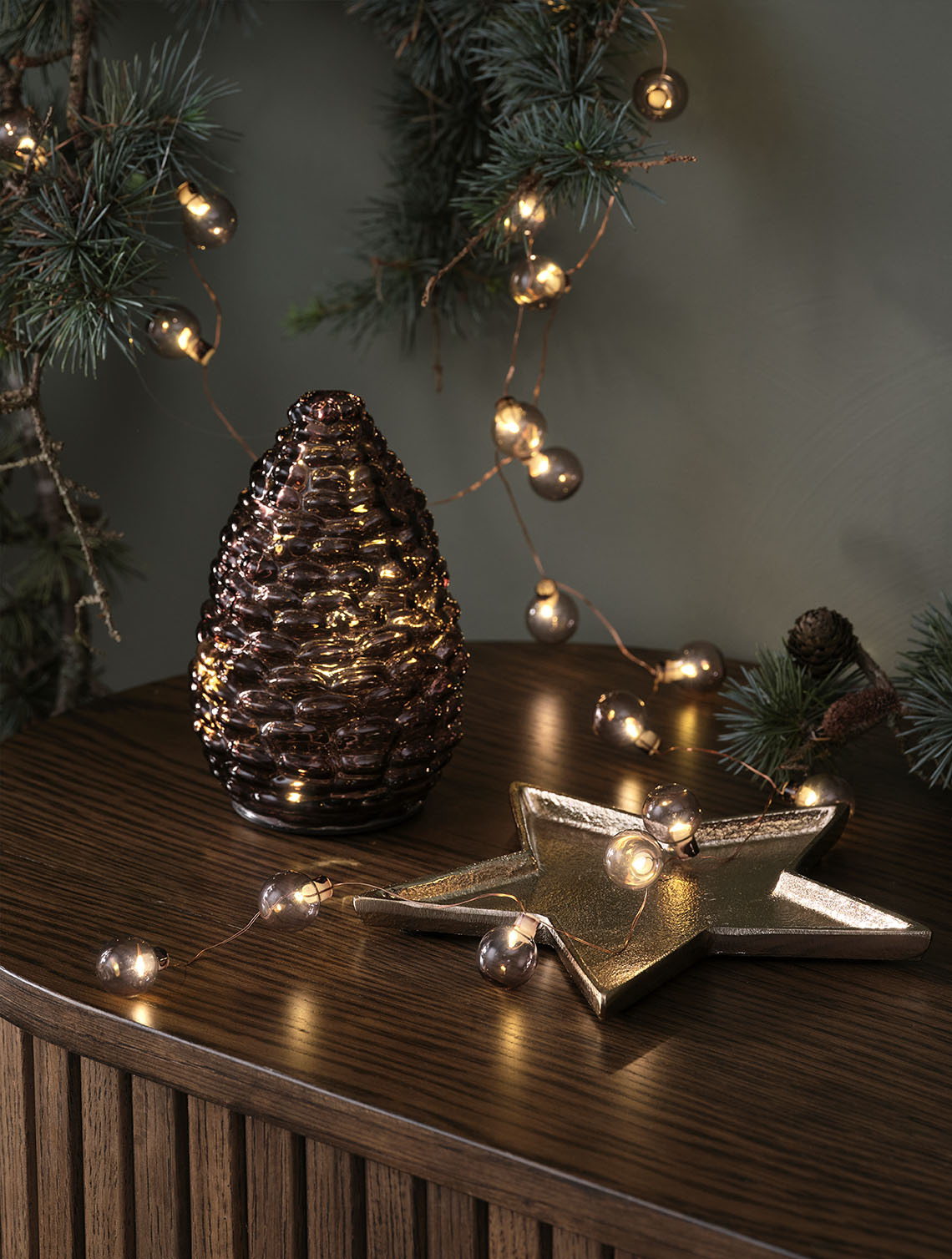 Decoratieve kerstverlichting, zoals een glazen dennenappel en een lichtslinger met gouden kerstballen op een dressoir.