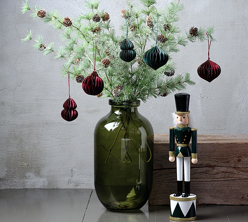 FELIX vaas gevuld met BURR takjes, versierd met PYROP versiering in groen en rood. Daarnaast een EUKLAS kerstbeeldje Groene glazen vaas gevuld met kunstmatige takjes met dennenappels, versierd met kerstversieringen in groen en rood. Daarnaast een kerstbeeldje