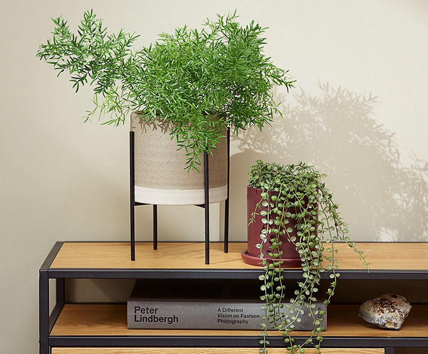 MANDRUP bloempot met poten en AXEL bloempot in paars met SVENN kunstplant Twee bloempotten op een modern TV-meubeld, een met een puntige plant en de ander met een hangende plant