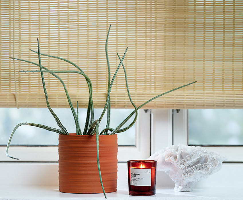 Bamboe rolgordijnen aan een raam met een bloempot, een geurkaars en een ornament op de vensterbank