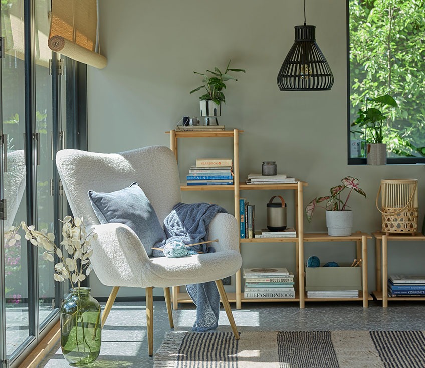 Off-white fauteuil bij het raam met een groene vaas op de vloer en een bamboe roomdivider op de achtergrond